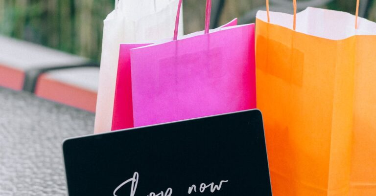 Laptop displaying 'Shop now' with vibrant shopping bags on glass table outdoors.